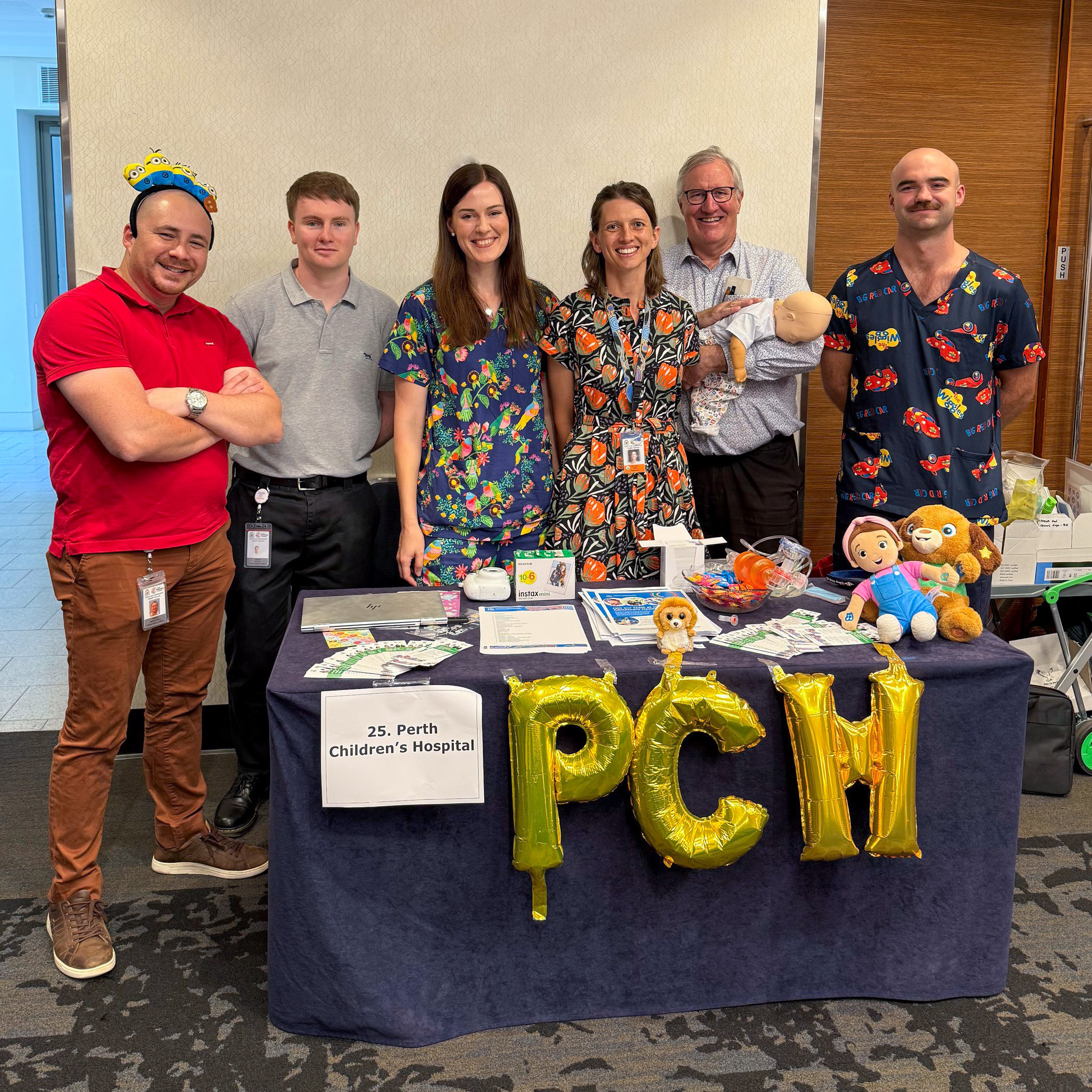 A group of six people stands behind an information table at an indoor event. The table is covered with a dark tablecloth displaying gold balloon letters spelling “PCH” and a sign that reads “Perth Children’s Hospital.” On the table are brochures, flyers, small giveaway items, a clear bowl of lollies, plush toy animals, and a mannequin baby used for demonstration. Several people wear patterned medical scrubs, while others wear casual or business-casual clothing. A wood-paneled wall and open foyer area are visible in the background.