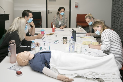 A group of people sitting around a table with a simulation manikin, taking part in an educational workshop